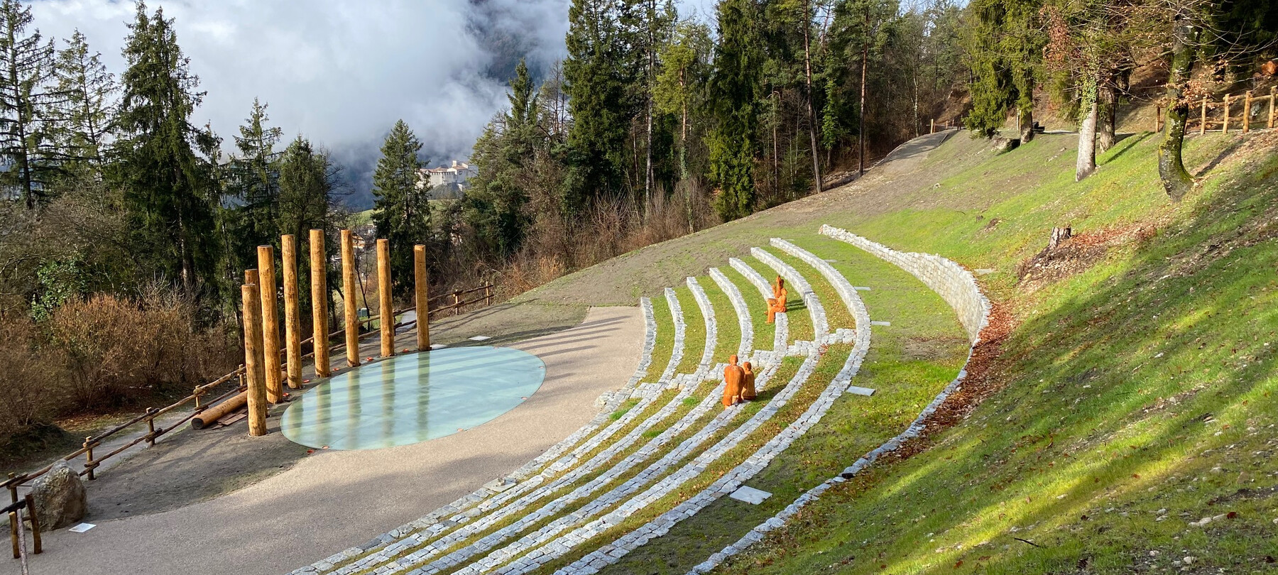 Art installation by Bosco Arte Stenico. A natural amphitheatre, in which stone steps follow the slope of the mountain. On the steps are four human figures, seated two by two. They are wooden statues, facing the arena of the amphitheatre, where there is a perfectly oval pool of water and, behind it, a fan of tall wooden columns. All around, pine, larch and beech trees. In the distance, white and grey clouds fill the sky and cover the mountain, which only peeps out occasionally where the clouds open up.