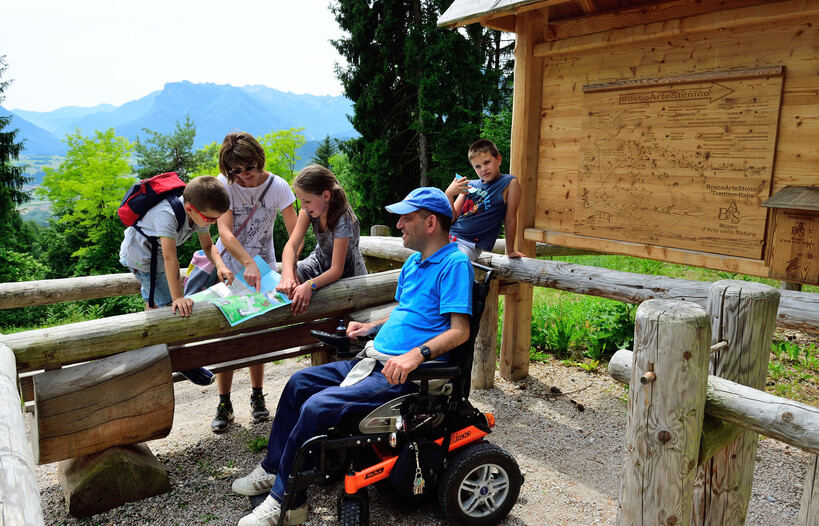 A small group of people, probably a family, consult a map before starting to explore Bosco Arte Stenico. In the image there are three children, two boys and a girl, a woman and a man in an electric wheelchair. Behind him, the forest path reproduced on a wooden panel, with an arrow indicating the direction to take to reach the beginning of the trail. It is a summer day, everyone is wearing light clothes and the sky is veiled by clouds that make the shadows soft.