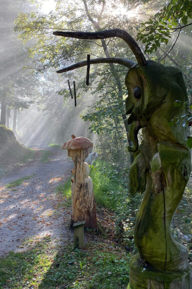Bosco Arte Stenico, on a sunny day when the rays create plays of light through the branches of the trees. On the side of the path, to the right of the image, two wooden statues. The one in the foreground, perhaps as tall as a man, could be a chimera: an owl's face, long horns resembling the antennae of an ant, its body seemingly wrapped in a tunic. The statue in the background, on the other hand, is smaller and resembles an apple core with a short stalk at the top.