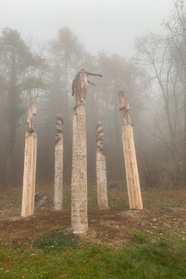 Five tall wooden columns stand out in the forest, on a misty day with little light. At the top of each column is a statue, also made of wood. In the statue in the foreground, two human figures, one male and one female, appear to be emerging from a wooden stump: their heads are leaning back and their left arm is raised, like a wing. In the statue on the right, two legs emerge from squared wooden stumps. The two statues in the background are faces with a strange headdress that spirals upwards: their mouths are open, they seem to be shouting. Finally, in the statue on the left, no human parts are visible: only wood, as if metamorphosis had not yet begun here. 