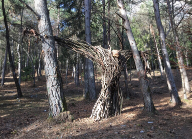 Art installation by Bosco Arte Stenico. The work of art is an intertwining of branches that evokes a human figure: its long arms are stretched out to embrace two trees, one on its right and one on its left. At the top, a wooden stump larger than the branches from which the work is made looks like the head of that half-man, half-tree figure.