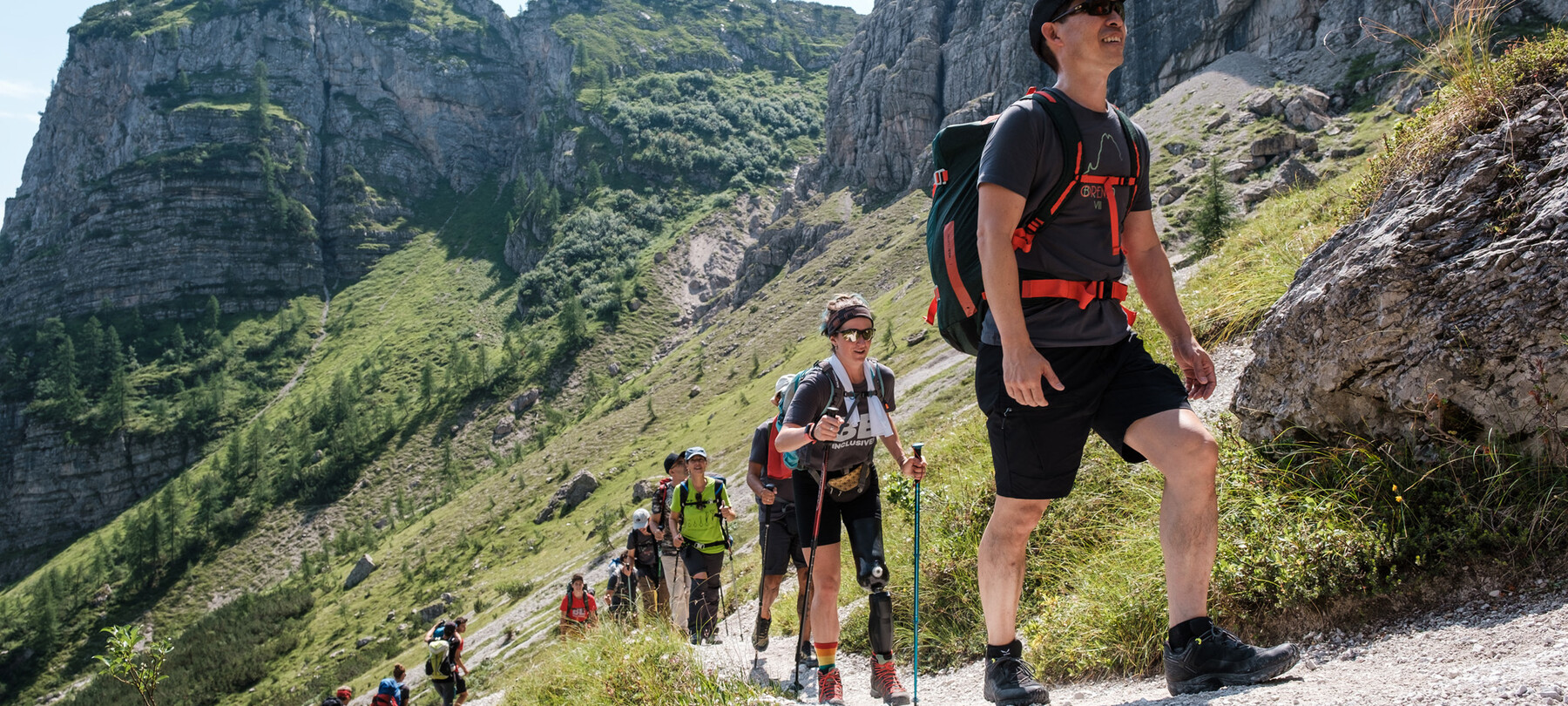 Un gruppo di persone, in fila, sta risalendo un sentiero sulle Dolomiti del Brenta. Seconda nella fila, una donna con una protesi alla gamba sinistra cammina con due bastoncini da trekking, supporto usato anche da altre persone del gruppo.