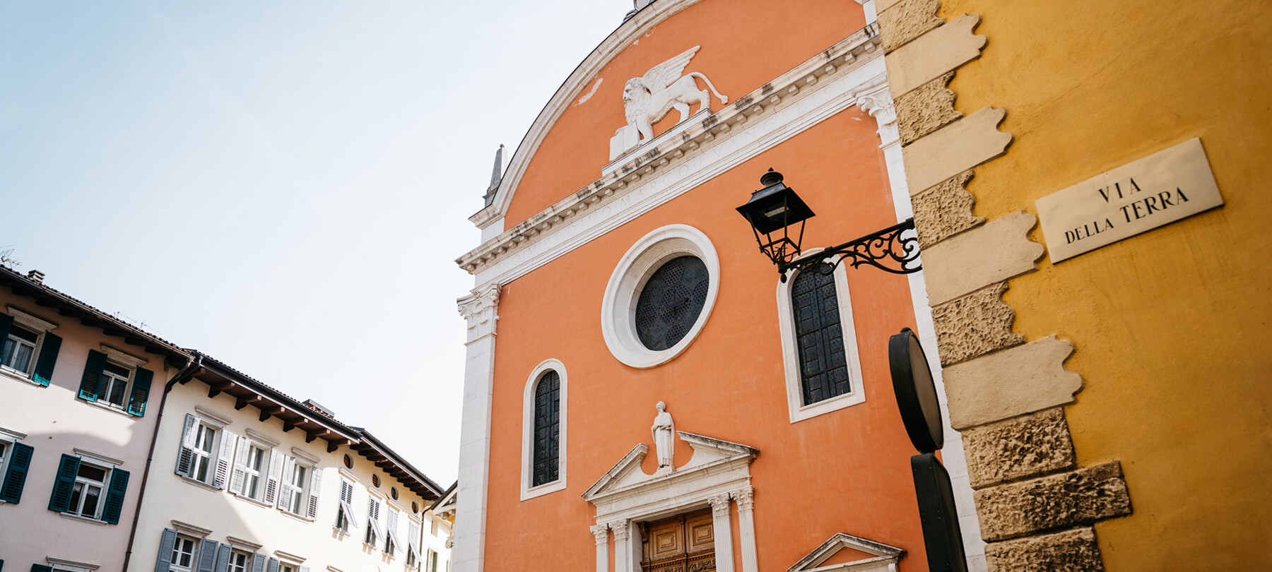 Facciata della Chiesa di San Marco, nel centro storico di Rovereto. Protagonisti dell’immagine sono i colori: l’arancione della chiesa, il giallo ocra del palazzo in primo piano, su cui si legge “Via della Terra”, il bianco e il rosa tenue degli edifici sullo sfondo. Anche se non è abitata da persone, è un’immagine vivace, gioiosa.