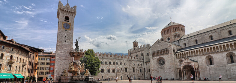 Piazza Duomo a Trento. Sullo sfondo, la Torre Civica, Palazzo Pretorio e la cattedrale di San Vigilio, Duomo della città. Al centro, la fontana del Nettuno. Dietro alla fontana svetta l’unico albero della piazza: un tiglio. La piazza, nell’immagine, è vissuta. Ci sono alcune persone sedute sui gradini della fontana, qualcuno sta scattando delle foto e un gruppetto è in posa davanti alla cattedrale per farsi fotografare. Tutti indossano abiti estivi. Il cielo azzurro è striato dal bianco di nuvole leggere.