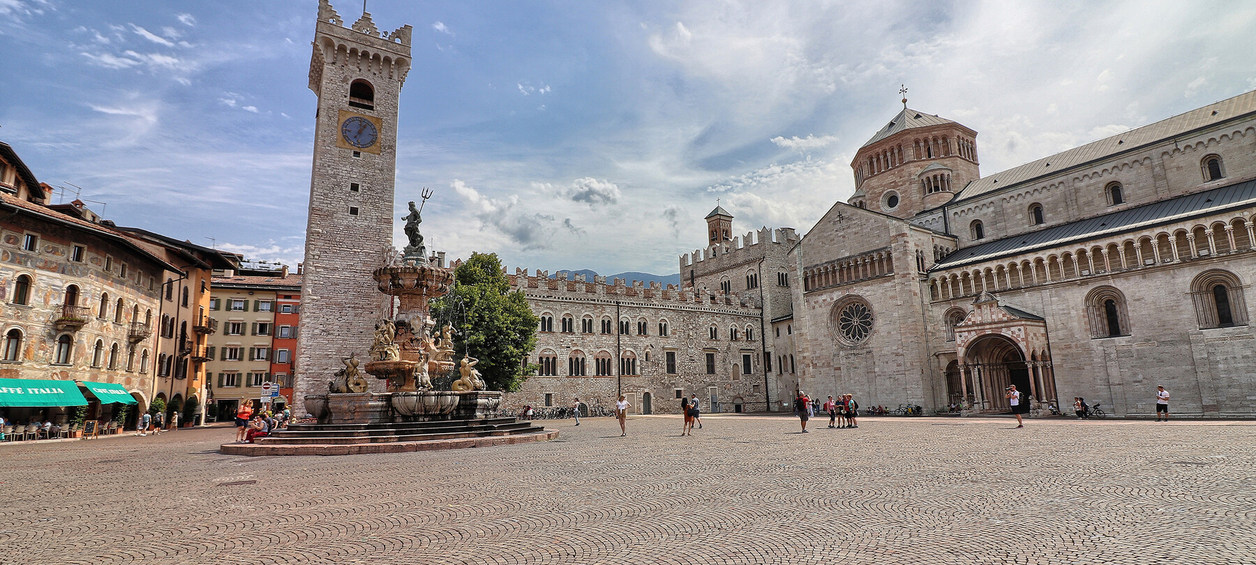 Piazza Duomo a Trento. Sullo sfondo, la Torre Civica, Palazzo Pretorio e la cattedrale di San Vigilio, Duomo della città. Al centro, la fontana del Nettuno. Dietro alla fontana svetta l’unico albero della piazza: un tiglio. La piazza, nell’immagine, è vissuta. Ci sono alcune persone sedute sui gradini della fontana, qualcuno sta scattando delle foto e un gruppetto è in posa davanti alla cattedrale per farsi fotografare. Tutti indossano abiti estivi. Il cielo azzurro è striato dal bianco di nuvole leggere.
