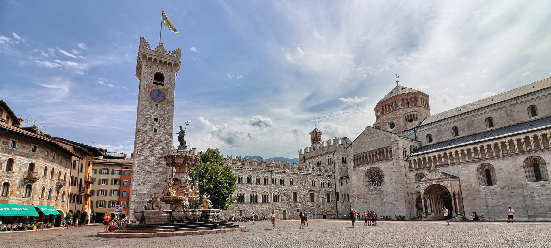 Piazza Duomo a Trento. Sullo sfondo, la Torre Civica, Palazzo Pretorio e la cattedrale di San Vigilio, Duomo della città. Al centro, la fontana del Nettuno. Dietro alla fontana svetta l’unico albero della piazza: un tiglio. La piazza, nell’immagine, è vissuta. Ci sono alcune persone sedute sui gradini della fontana, qualcuno sta scattando delle foto e un gruppetto è in posa davanti alla cattedrale per farsi fotografare. Tutti indossano abiti estivi. Il cielo azzurro è striato dal bianco di nuvole leggere.