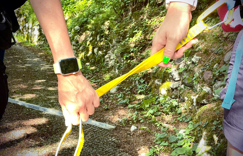 Two hands hold a yellow strap that is used to facilitate mountain hikes for blind or visually impaired people. On one side, the strap is tied to a carabiner attached to the backpack of the person leading the walk. On the other side, the hand gripping it is the blind or visually impaired person's. In the background, the road they are walking along: between the brown of the path, the grey of the stones and the green of the leaves, the rays of the sun create small plays of light and reflections. 