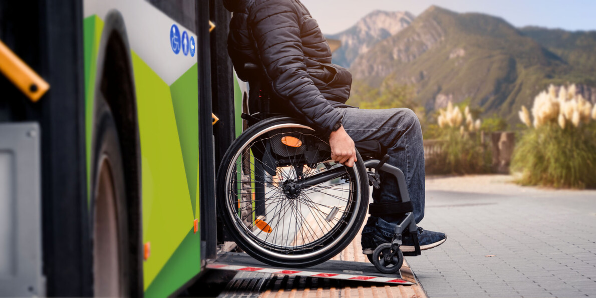 Un uomo in sedia a rotelle scende da un autobus di linea urbana di Trentino Trasporti utilizzando la rampa che permette alle carrozzine di accedere alla vettura. Sullo sfondo, le montagne illuminate da una luce calda e morbida.