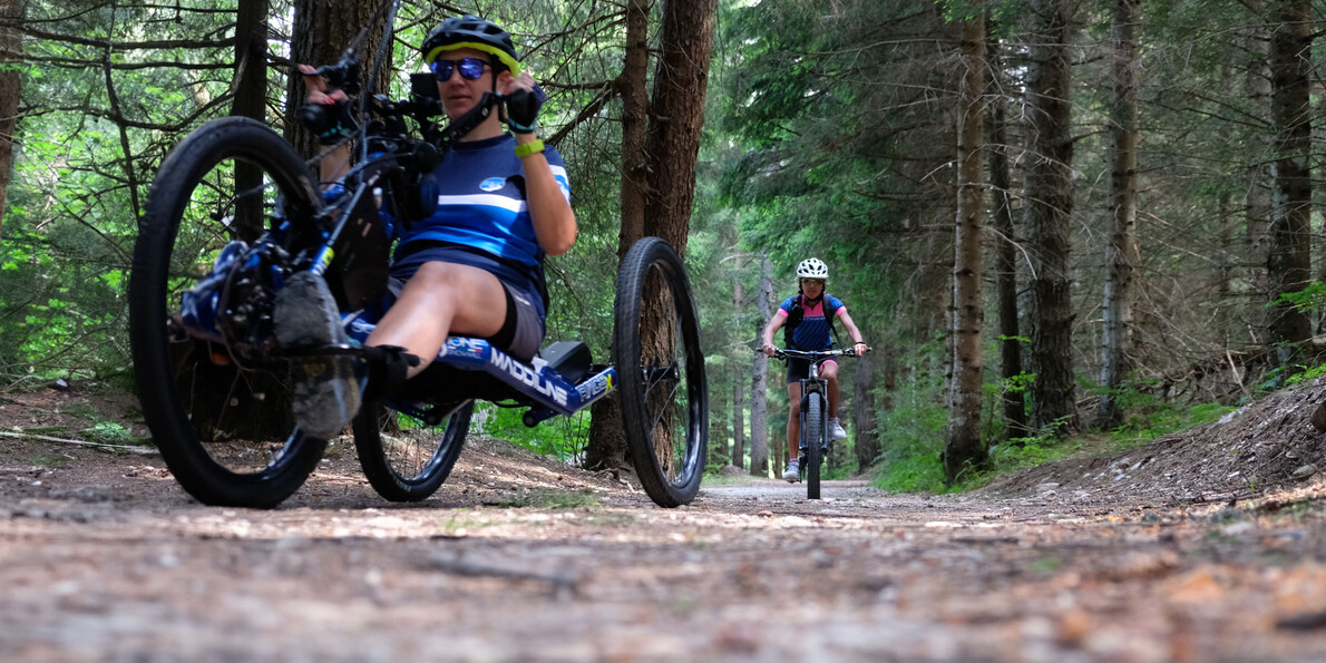 Two people are biking along a forest path. The person in the background rides a classic bicycle, while the one in the foreground rides a handbike, operated by the upper body.