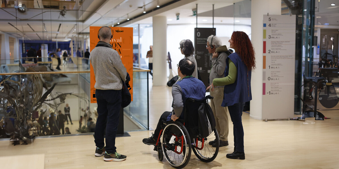A group of people visit one of the locations of the Festival of Economics. Among them is a man in a wheelchair: the space has no architectural barriers and is also accessible to those with mobility disabilities. All the people are wearing earphones: they are probably enjoying audio content related to the Festival.