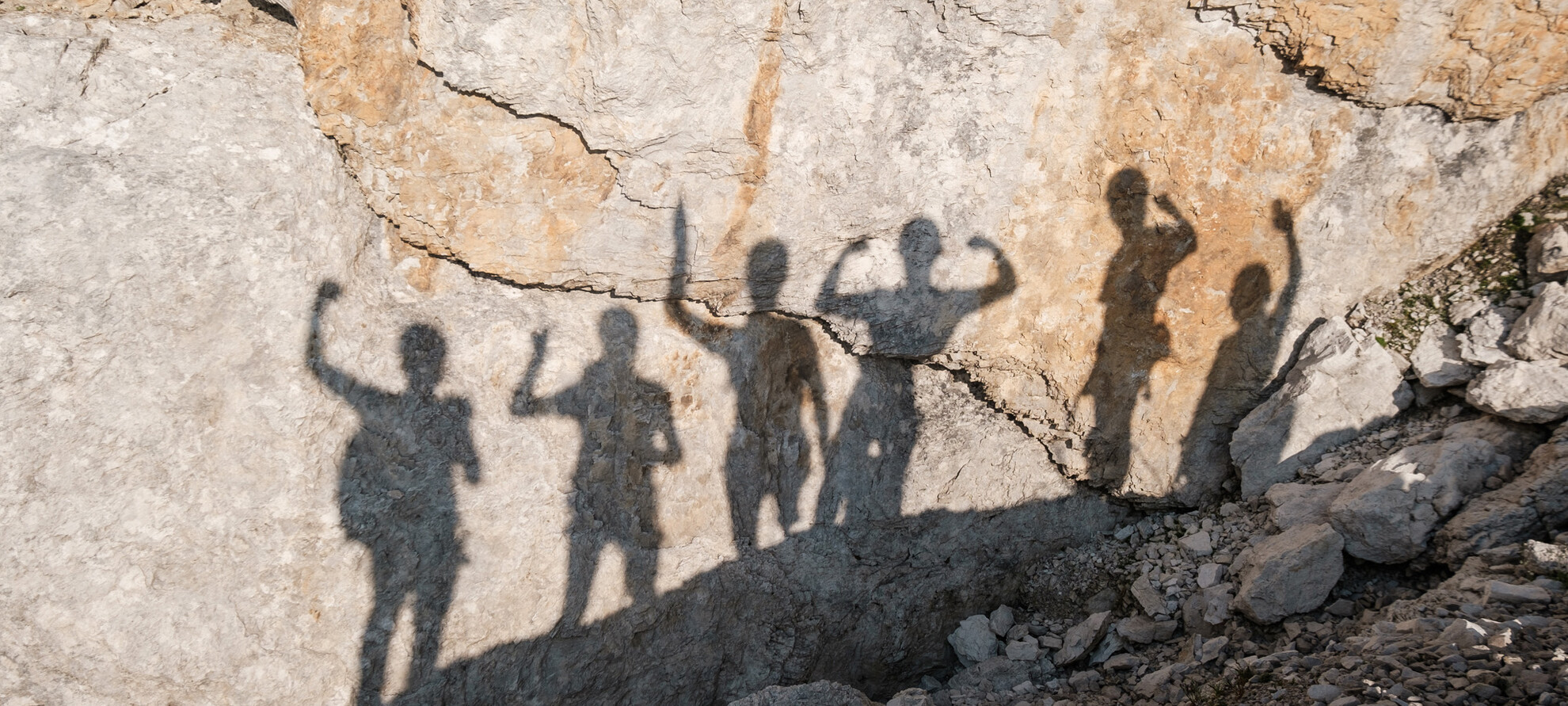 Sei alpinisti esultano per aver raggiunto la cima. Nella foto ci sono solo le loro ombre sulla superficie rocciosa della montagna. Braccia sollevate e segni di vittoria: la soddisfazione di avercela fatta, insieme.