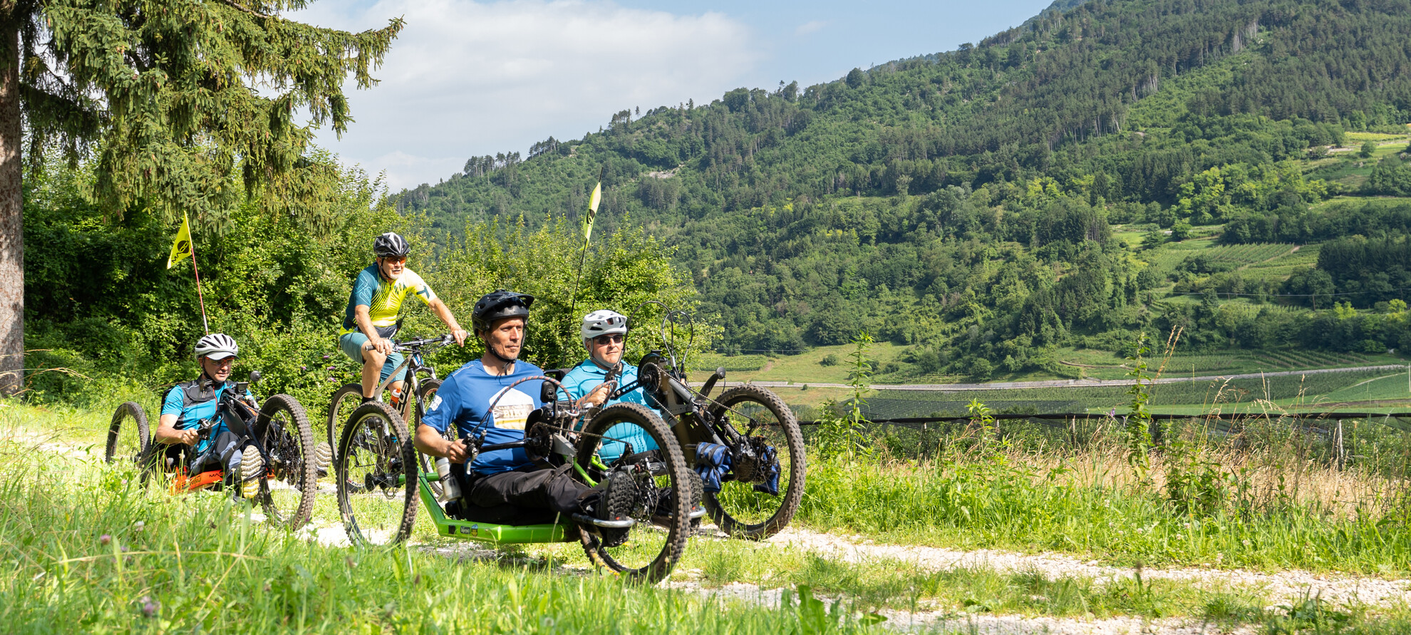 Siamo sull’Altopiano della Vigolana. È una bella giornata estiva, luminosa. Quattro turisti, amanti dell’attività outdoor, fanno biking seguendo un sentiero sterrato: tre di loro guidano una handbike. Insieme, si godono il sole sulla pelle, l’aria fresca e il verde dei boschi che li circonda. | © ©Trentino Marketing_Gianluca Prati_Accessibilita_Vigolana_2024_00006