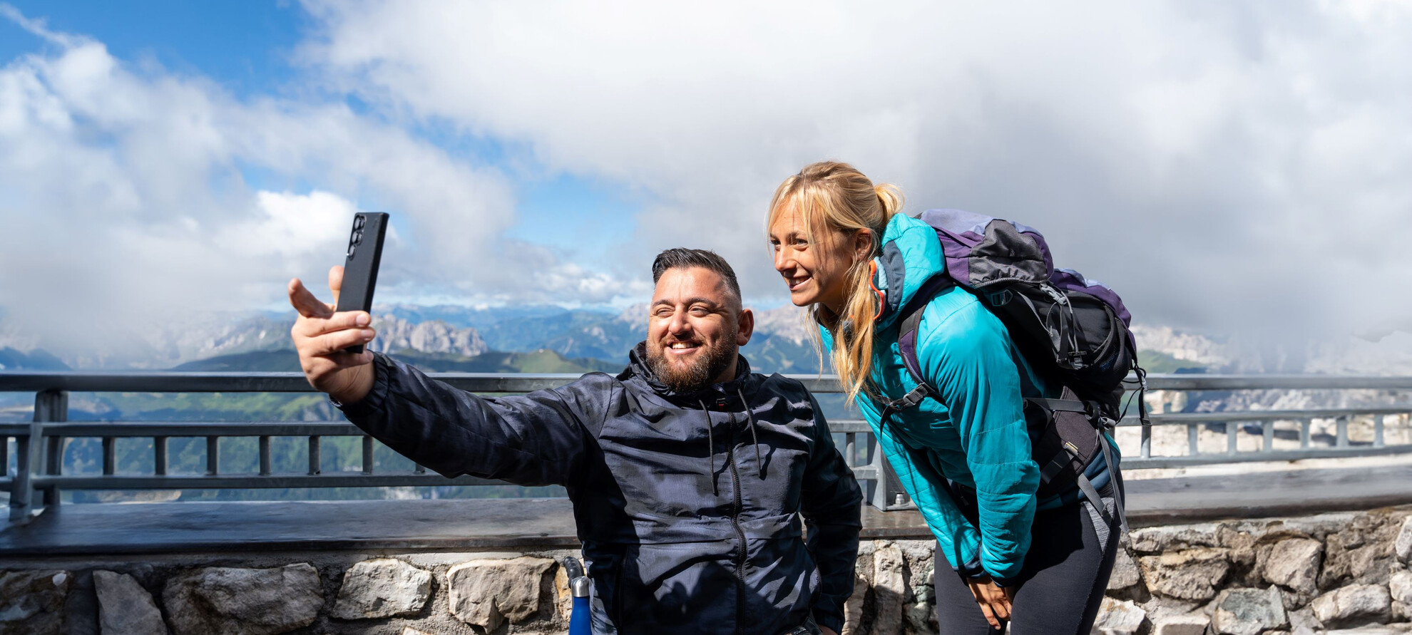 Un uomo e una donna in abbigliamento sportivo si fanno una foto dal punto panoramico del Sass Pordoi. Dietro di loro, sullo sfondo, le cime delle Dolomiti sono coperte dalle nuvole che lasciano intravedere l’azzurro del cielo.  | © ©Trentino Marketing_Gianluca Prati_Accessibilita_Val di Fassa_2024_00024