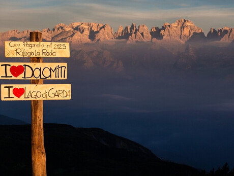 L'immagine  mostra le Dolomiti di Brenta all'alba e davanti un cartello con le scritte "Cima Paganella 20125 -Rifugio La Roda" e I love Dolomiti e lago di Garda