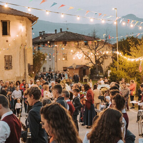 A warm and festive atmosphere fills the streets of Zambana Vecchia during Antiche Terre. The band plays surrounded by people in vintage clothing, under string lights and colorful flags. The community gathers to celebrate tradition and memory.