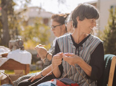 A group of women are knitting along the main street of Zambana Vecchia. They have many fabrics in different colors and enjoy showing people what they’re making. Behind them, there’s a small table with an old sewing machine and other antique objects.
