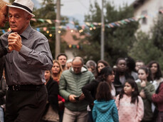 A couple dances tenderly in the town square during the festival, surrounded by onlookers of all ages. String lights and colorful flags decorate the scene. The moment conveys a sense of community, warmth, and togetherness.