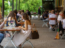 Many people seated at tables are enjoying a summer aperitif under the veranda of the Endrizzi Winery. The atmosphere is elegant yet welcoming. String lights on the roof create a warm ambiance. Around the veranda, a vineyard glows in vibrant green. Glasses of wine are served with delicious appetizers.