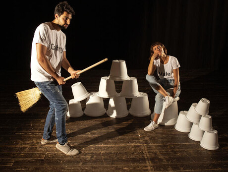 L'immagine mostra gli attori durante una scena dello spettacolo: lui che cavalca una scopa, lei seduta su alcuni vasi bianchi