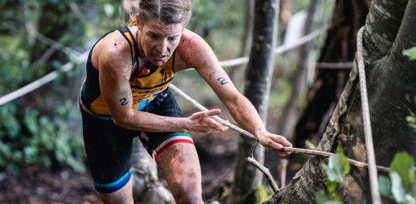 An X-Terra participant climbs up from a steep forest clinging to a rope.