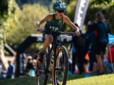A child pedals his bike with determination on the beach of Lake Molveno. He wears a helmet, sunglasses and a green uniform, with ITA written on his chest.