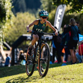 A child pedals his bike with determination on the beach of Lake Molveno. He wears a helmet, sunglasses and a green uniform, with ITA written on his chest.