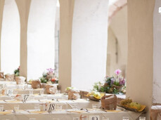 Many wooden tables set with white tablecloths under the cloister of the Augustinian convent of San Michele all’Adige. Beside the tables, there is a wooden decoration with apples and grapes, while beneath the arches of the portico, colorful and elegant floral arrangements are displayed.