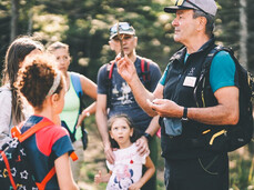 Una guida escursionistica, durante un'uscita nel bosco a Fai della Paganella, mostra un rametto a un gruppo di partecipanti, adulti e bambini. Le persone sono radunate intorno a lui e ascoltano con attenzione la spiegazione sulla natura.