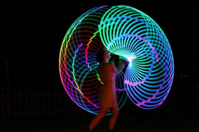 Nell’immagine si vede una persona che si esibisce di notte all’aperto, creando con il movimento grandi cerchi colorati e luminosi che formano un effetto a spirale nel buio. I colori accesi — blu, verde, rosso e giallo — illuminano la scena come un disegno di luce in movimento.