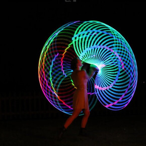 Nell’immagine si vede una persona che si esibisce di notte all’aperto, creando con il movimento grandi cerchi colorati e luminosi che formano un effetto a spirale nel buio. I colori accesi — blu, verde, rosso e giallo — illuminano la scena come un disegno di luce in movimento.