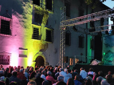 The image shows an audience of about 200 people seated, waiting for the show to begin. The chairs are arranged in the courtyard of a 19th-century building, illuminated by yellow and pink lights. On the stage, there are structures that are likely part of the upcoming performance. The people are smiling and eagerly anticipating the start of the show.