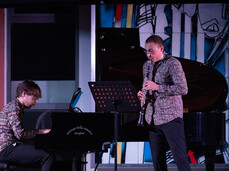 This photo shows two musicians performing on stage: a pianist seated at the piano and a clarinetist standing next to a music stand. They wear similarly patterned shirts and play with deep concentration. In the background, there is an abstract artwork, and the piano lid is open
