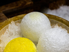 La foto inquadra un secchio di legno appoggiato ad una seduta di una sauna in legno. Al suo interno ci sono tre palle di ghiaccio. Su una palla di ghiaccio è stato già versato l'olio essenziale profumato. Si tratta delle palle di ghiaccio utilizzate durante i riti di sauna 'Aufguss'