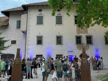 A group of people gathers in front of a historic building with walls illuminated by blue lights. The atmosphere is friendly and sociable; it is a cultural and food-and-wine event. Two metal structures bearing the word "MEZZOCORONA" mark the entrance.