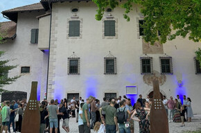 A group of people gathers in front of a historic building with walls illuminated by blue lights. The atmosphere is friendly and sociable; it is a cultural and food-and-wine event. Two metal structures bearing the word "MEZZOCORONA" mark the entrance.