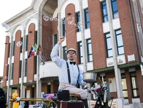 La foto ritrae un artista di strada, in arte "Bolle Girovaghe", intento a creare bolle di sapone con una bacchetta, davanti a un edificio pubblico. L’atmosfera è vivace e cattura l’attenzione con il suo stile rétro e il carretto ricco di strumenti.