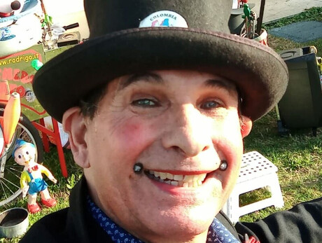 La foto mostra Clown Rodrigo, sorridente e vestito in stile circense con un cappello a cilindro, in un ambiente all'aperto. Alle sue spalle, oggetti colorati e giocattoli contribuiscono a creare un’atmosfera allegra e vivace, ideale per l’intrattenimento.