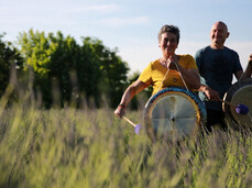 The photo shows two musicians walking in nature. They are two gong players and they hold the gong in one hand and the stick to play it in the other. Their faces communicate passion for what they do and serenity. They are in the tall grass, the sun shines low behind them.