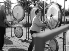 The photo shows two musicians walking in nature. They are two gong players and they hold the gong in one hand and the stick to play it in the other. Their faces communicate passion for what they do and serenity. They are in the tall grass, the sun shines low behind them.