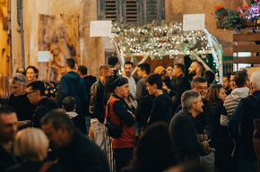 The photo shows a lively outdoor party in a historic village, illuminated by hanging lights and floral decorations. People are chatting and enjoying the convivial atmosphere, with stalls and decorated arches. The ancient walls and wooden shutters add rustic charm.