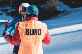 Uno sciatore non vedente, riconoscibile dalla pettorina arancione con la scritta "BLIND", viene guidato da un accompagnatore su una pista da sci innevata.