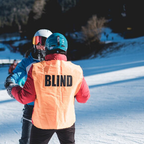 Uno sciatore non vedente, riconoscibile dalla pettorina arancione con la scritta "BLIND", viene guidato da un accompagnatore su una pista da sci innevata.