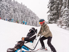 A ski instructor goes down a slope carrying a monoski, an aid for people with disabilities. It is snowing heavily and the falling snow partly covers the ski instructor and the person on the monoski.