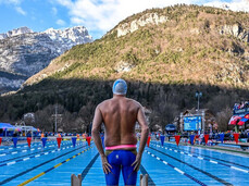 A swimmer, seen from behind, stands on starting block number 6, overlooking an outdoor pool set for ice swimming. The athlete, wearing swim briefs and a cap, prepares for the race in an alpine setting, with imposing mountains surrounding the facility.