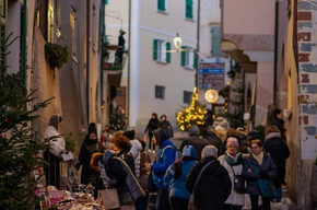 L'immagine mostra una delle vie del borgo di Faedo abbellita da addobbi natalizi. Lungo la via ci sono diverse persone che passeggiano e guardano le bancarelle del mercatino di Natale. 
