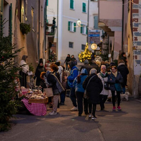 L'immagine mostra una delle vie del borgo di Faedo abbellita da addobbi natalizi. Lungo la via ci sono diverse persone che passeggiano e guardano le bancarelle del mercatino di Natale. 