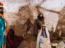 The photo shows the Nativity scene depicted with life-sized statues, featuring Joseph, Mary, and the Baby Jesus. Behind them are a cow and a Wise Man. The entire scene is set inside a vaulted passageway in the village of Faedo.