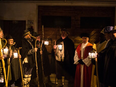 The photo shows a group of people dressed in traditional costumes taking part in the Canto della Stella in Faedo. Some hold staffs and lanterns, creating an evocative atmosphere. The scene takes place in the evening, lit by the warm glow of the lamps.