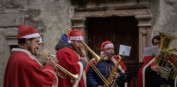 L'immagine ritrae 4 uomini vestiti con cappello e mantello di Babbo Natale suonano trombe e tromboni intonando canti natalizi