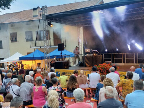 The image shows an outdoor community event with a stage where a person is operating equipment, possibly a DJ or presenter. A crowd of people, mostly middle-aged and older, watches the performance while seated or standing in an open area with a reddish floor. The setting appears to be in a small town or village with mountains visible in the background, and there are some market tents and colorful decorations visible, suggesting it might be a local festival or cultural celebration.